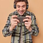 optimistic-addicted-man-gamer-plays-video-games-smartphone-stares-screen-cellular-feels-glad-wears-wireless-headphones-ears-dressed-checkered-shirt-isolated-beige-background (1)